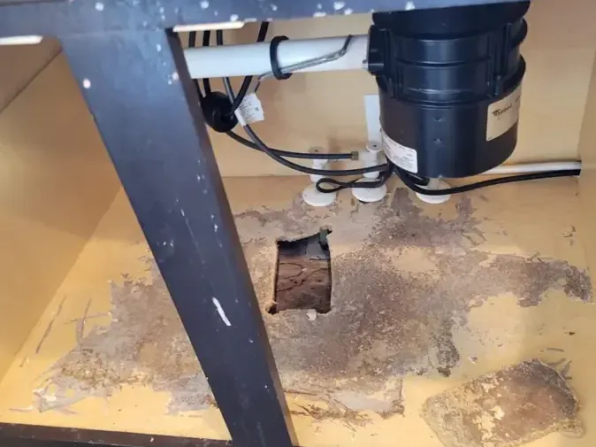A water-damaged sink cabinet floor with an access hole cut below the plumbing