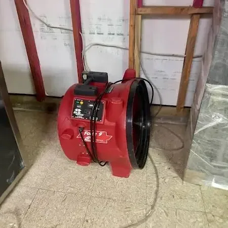 Plastic sheeting hung beside an opened lower wall while a red air mover dries the room after a leak